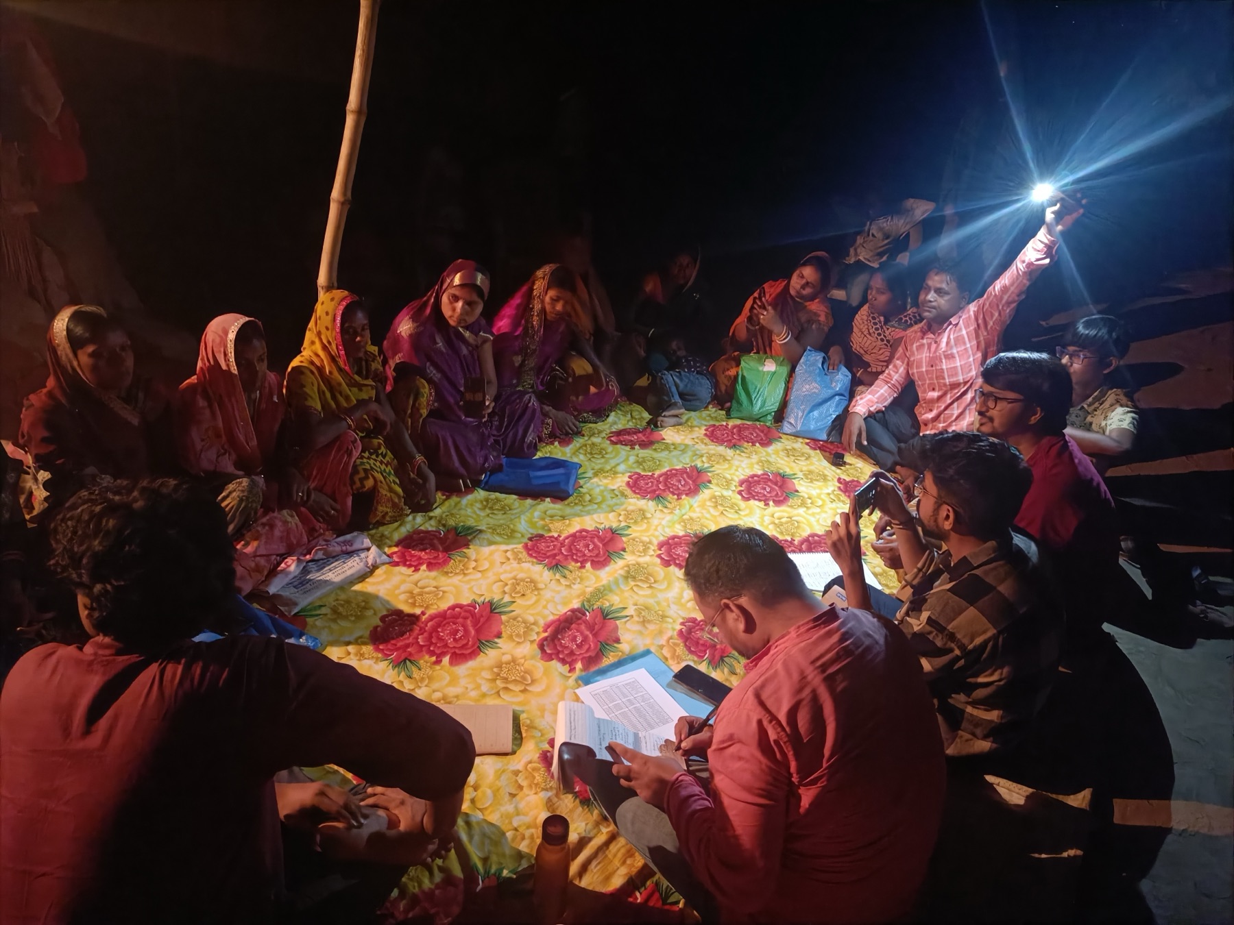 Night-time community meeting with women in a village, lit by flashlight